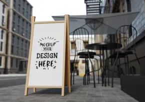 A sidewalk sign mockup advertising design services next to outdoor seating in an urban setting. - PSD Mockup