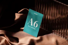 An A6-sized teal book titled "The A6" stands upright on a shiny, wavy satin fabric under a warm, dim light, serving as an ideal mockup. - PSD Mockup