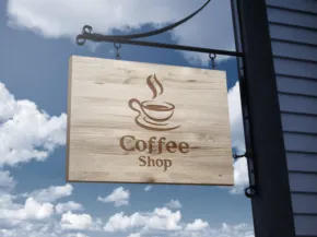 A wooden sign mockup with "coffee shop" and a coffee cup symbol hanging outside under a blue sky with clouds. - PSD Mockup