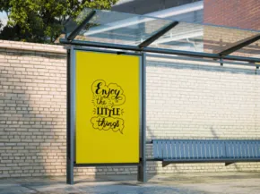 A yellow poster on a bus stop shelter reads "Enjoy the Little Things" in black script. The shelter, which could serve as an excellent mockup template for advertisements, features a metal bench and a glass roof, set against a brick wall backdrop. - PSD Mockup