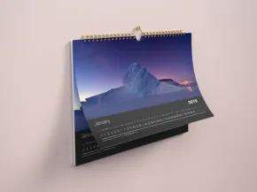 Desk calendar mockup open to a template page showing a mountain landscape at sunset, with text and dates visible. - PSD Mockup