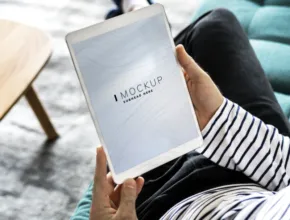 Person sitting on a couch, holding a tablet with a screen displaying the word "mockup". - PSD Mockup