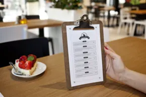 A person holding a template on a clipboard in a cafe, with a dessert plate containing a tart topped with fruits on the table. - PSD Mockup