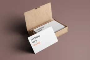 A small open cardboard box with a mockup of business cards inside, one card partially pulled out; placed on a solid beige surface. - PSD Mockup