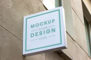 A square hanging sign on a building wall, displaying the text "mockup your design here" in a clean, modern font. - PSD Mockup