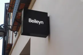A black square sign template with the word "beilwyn" in white letters, mounted on a light-colored wall under a roof edge. - PSD Mockup
