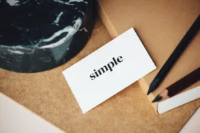 A mockup featuring a card with the word "simple" next to a black stone and pencils on a brown textured paper background. - PSD Mockup