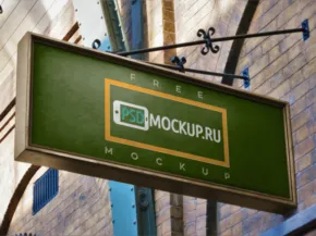 A hanging rectangular green and white sign with the text "template mockup" displayed on a sunny day, with a brick building in the background. - PSD Mockup