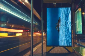 A digital billboard template displaying a surfer riding a large wave at night on a city street, with light trails from passing vehicles. - PSD Mockup