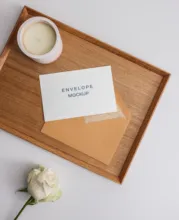 A wooden tray with a beige envelope labeled "events welcome," serving as a mockup, a small cup of cream, and a white rose on a white background. - PSD Mockup