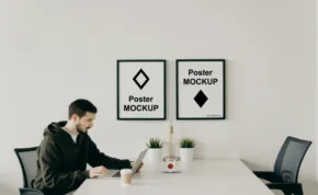 A man sitting at a table using a smartphone with a laptop beside him, under two framed mockup posters on a white wall. - PSD Mockup