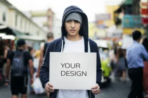 Young person holding a sign with "mockup template" written on it, standing in a crowded street. - PSD Mockup