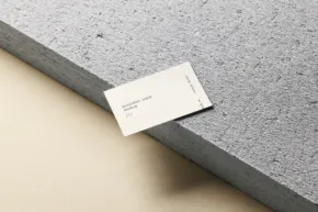 A business card mockup with a minimalistic design rests on the edge of a gray stone slab against a beige background. - PSD Mockup