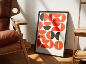 A framed abstract artwork with red and black circles leaning against a wall in a sunny room with wooden furniture serves as an ideal mockup. - PSD Mockup