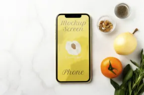 Smartphone with a yellow template screen lying on a white surface next to leaves and spices. - PSD Mockup