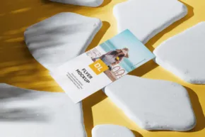 Brochure mockup featuring a person's photo lying among white, irregularly shaped stones on a yellow surface with sunlight and shadows. - PSD Mockup