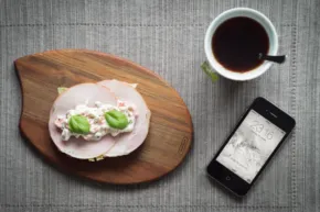 A wooden board with an open-faced sandwich, a cup of black coffee, and a smartphone displaying a map on a mockup template, all set on a gray textured tablecloth. - PSD Mockup