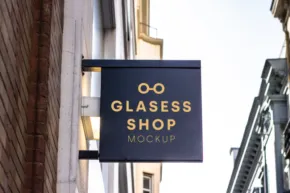 A square sign template hanging on a building's facade that reads "glasses shop mockup" with an icon of glasses above the text. - PSD Mockup