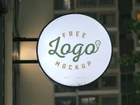 A circular illuminated sign displaying the template "free logo mockup" mounted on a storefront window, with blurred buildings in the background. - PSD Mockup