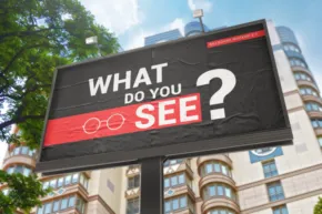 A digital billboard mockup displaying the question "what do you see?" in white letters on a red background, located in a city setting with modern buildings and trees under a clear blue sky. - PSD Mockup
