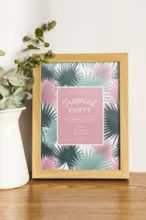 A framed tropical party print *template* on a table next to a potted plant. - PSD Mockup