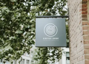 A sign for a coffee shop is hanging on the side of a brick building, serving as a mockup. - PSD Mockup